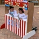 Trois jeunes enfants jouant avec des marionnettes derrière un théâtre de marionnettes à rayures rouges et blanches.