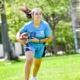 Une personne aux longs cheveux noirs et en tenue de sport court sur un terrain herbeux en portant un ballon de football.