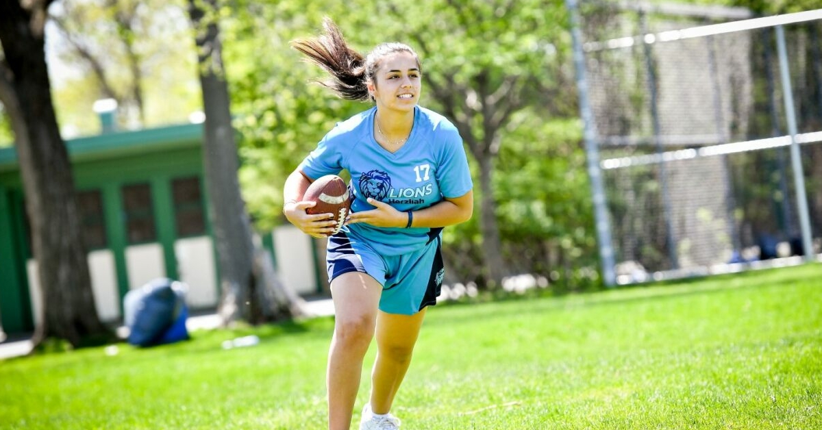 Une personne aux longs cheveux noirs et en tenue de sport court sur un terrain herbeux en portant un ballon de football.