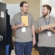 Deux personnes portant des t-shirts assortis marchent et parlent. L'une porte une boîte, l'autre une enveloppe.