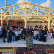 Large group of people, seated and standing in front of a partially built outdoor structure, listening someone giving a speech.