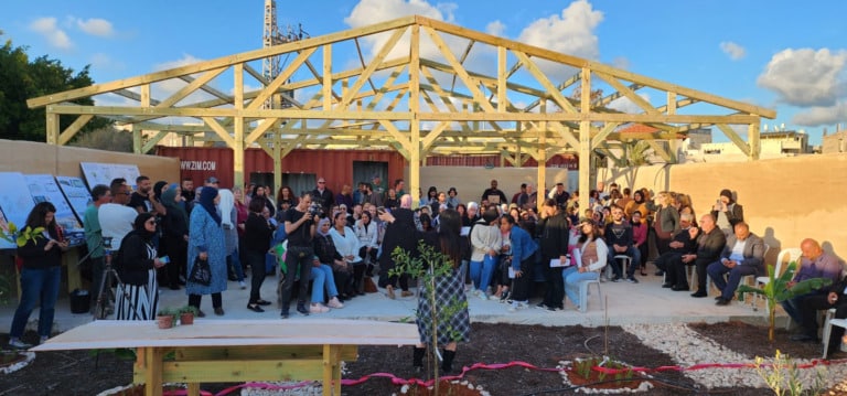Large group of people, seated and standing in front of a partially built outdoor structure, listening someone giving a speech.