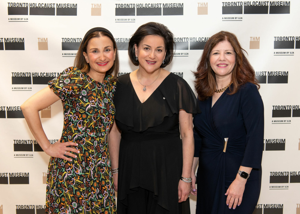 Three people in formal dresses in front of a wall with Toronto Holocaust Museum logo.