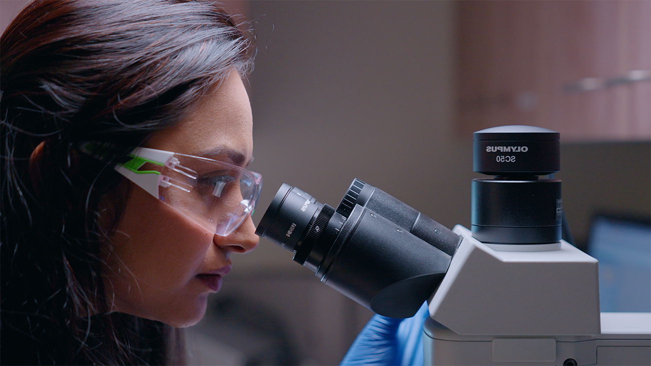 A person with long dark hair wearing blue gloves and protective goggles looking into a microscope.