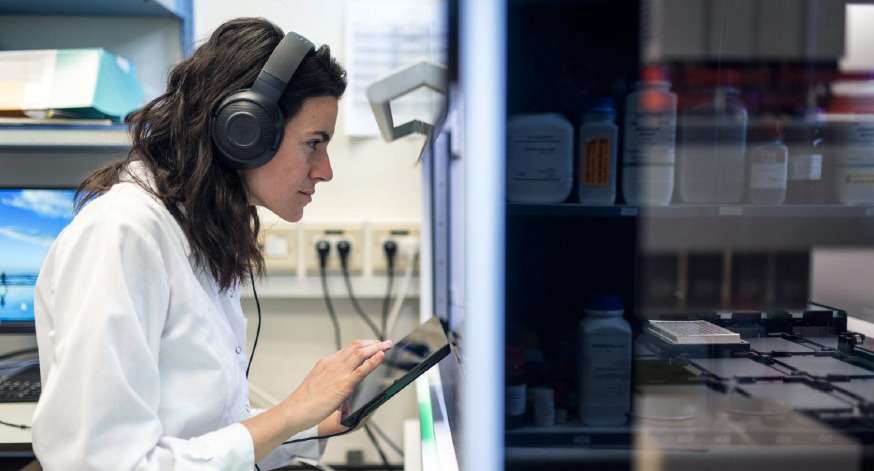 A person in a lab coat wearing headphones and looking at a tablet.