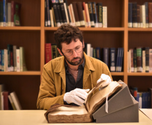 A person with glasses and white gloves reading a large, old book in a library.