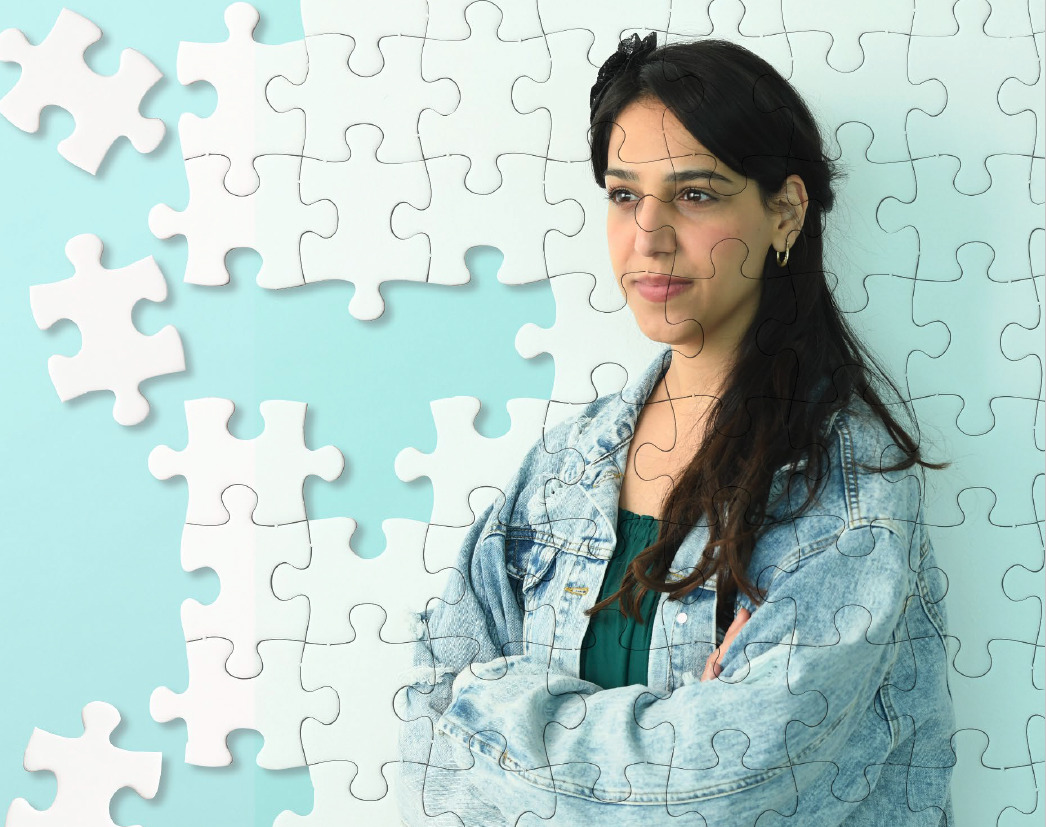 A person with long hair an a denim jacket crossing their arms and smiling. The image appears as a puzzle, a few pieces have yet to be secured.