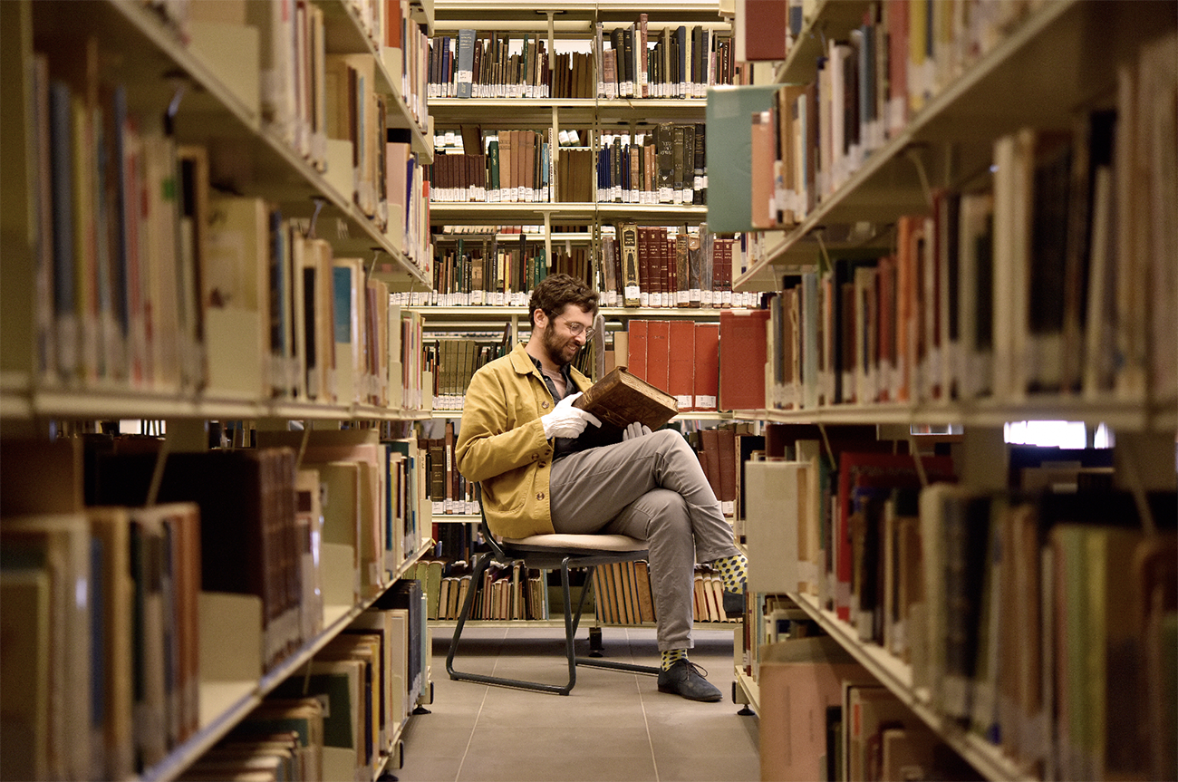A person sitting on a chair reading a book between the tall shelves of a library.