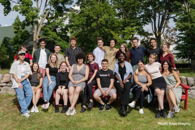 A large group of young adults outdoors. There is a row of people is sitting on red chairs while others stand behind them.