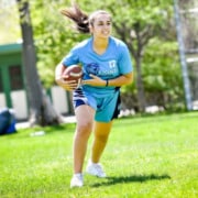 A person with long dark hair and athletic clothing runs on a grassy field carrying a football.
