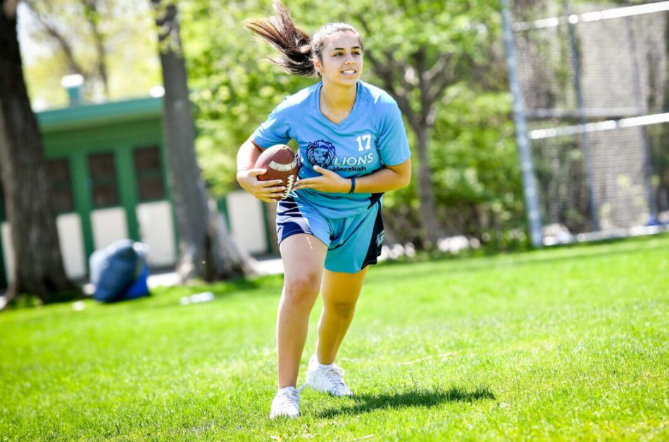 A person with long dark hair and athletic clothing runs on a grassy field carrying a football.