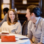 Two people sitting and talking at a large banquet table. There are two notebooks on the table.