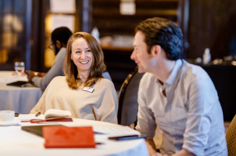 Two people sitting and talking at a large banquet table. There are two notebooks on the table.