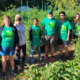 Six individuals standing in a very green garden wearing green tshirts.