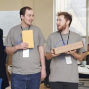 Two people wearing matching tshirts walking and talking. One is carrying a box, and the other carries an envelope.