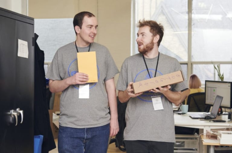 Two people wearing matching tshirts walking and talking. One is carrying a box, and the other carries an envelope.