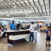 A local library scene. One person checking out a book at the desk with the librarian, while a person and their child stand nearby.