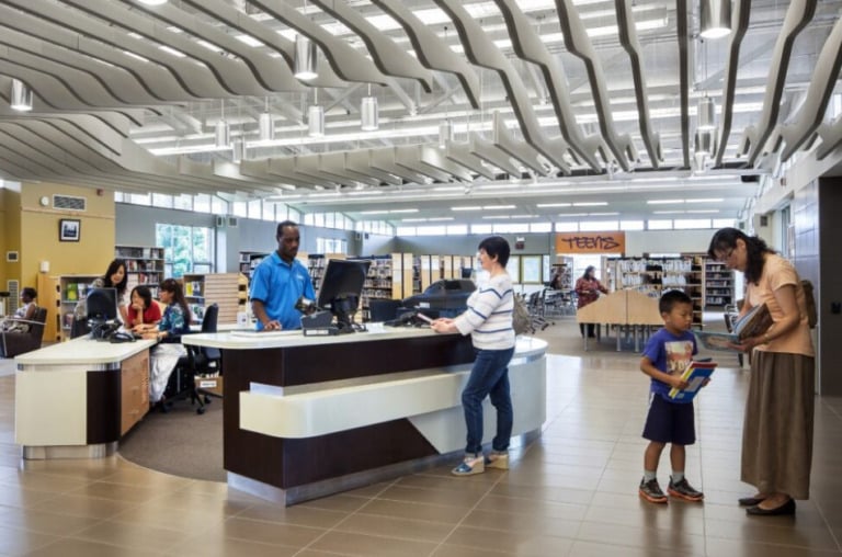 A local library scene. One person checking out a book at the desk with the librarian, while a person and their child stand nearby.