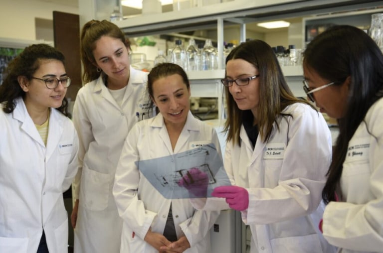Five scientists in a lab. One is wearing pink gloves and holds up a sheet of paper while the other three look on.