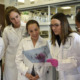 Five scientists in a lab. One is wearing pink gloves and holds up a sheet of paper while the other three look on.