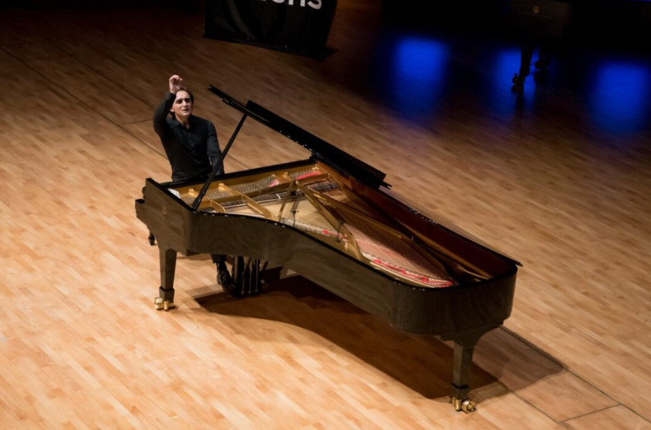 A person sits on a stage, and they are alone at a grand piano. One of their arms is raised as if to play.