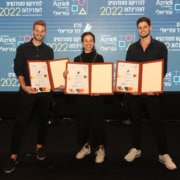 Three young adults stand in front of a backdrop with the Azrieli Foundation logo. They each hold up a portfolio with an award.