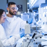Two scientists in a lab. Both are wearing goggles, white lab coats and blue gloves.