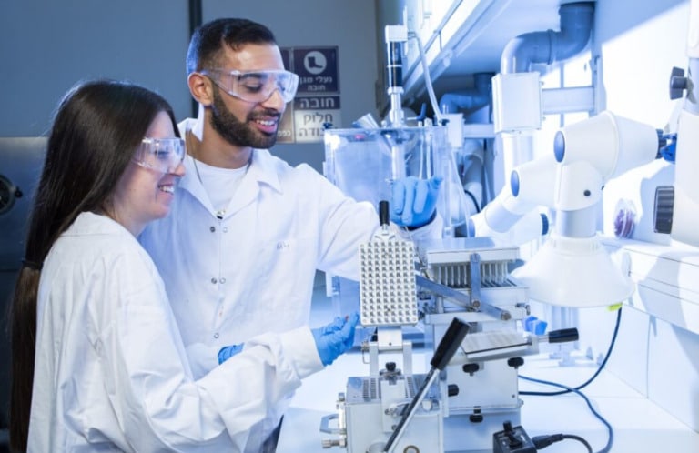 Two scientists in a lab. Both are wearing goggles, white lab coats and blue gloves.