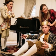 Three people in old English costumes. One sits confused on the floor, another tries to help them, a third looks on with concern.