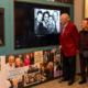 Two older adults stand in front of a museum exhibit screen, projecting a black and white photo.