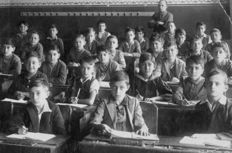 A black and white photo of young students at their desks in a crowded classroom.