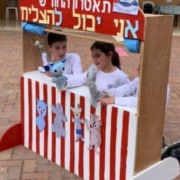 Three young children playing with puppets behind a red and white striped puppet theatre.