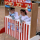 Three young children playing with puppets behind a red and white striped puppet theatre.