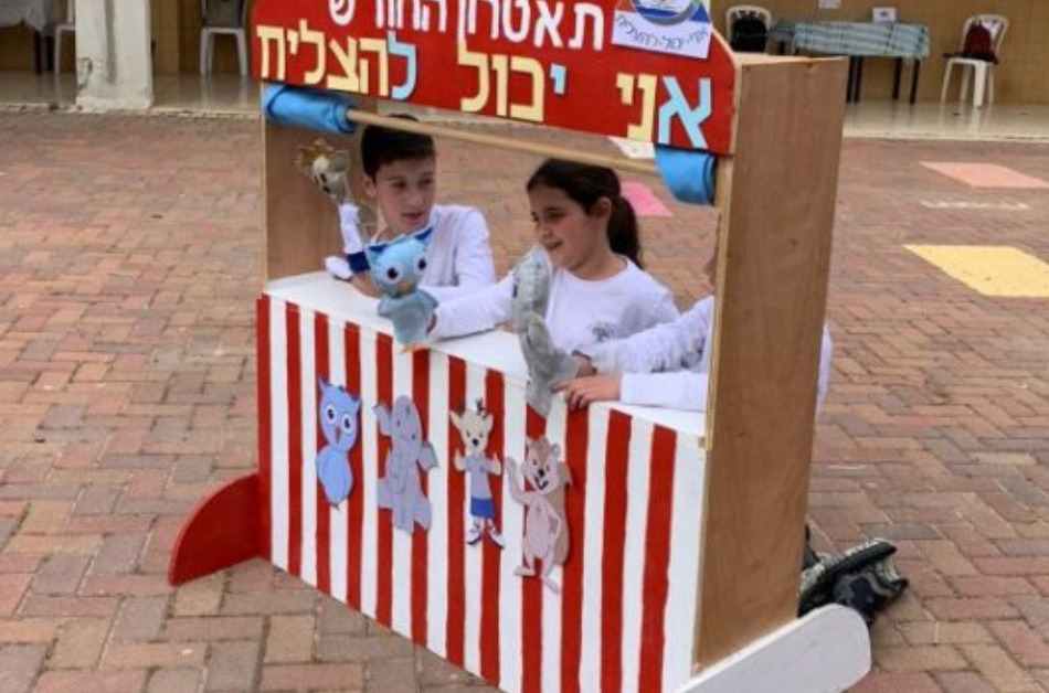 Three young children playing with puppets behind a red and white striped puppet theatre.